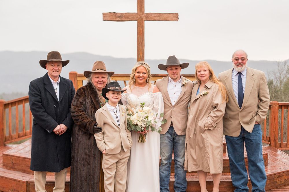 A group gathers for a wedding photo in front of a wooden cross on an outdoor deck overlooking mountains in the background.