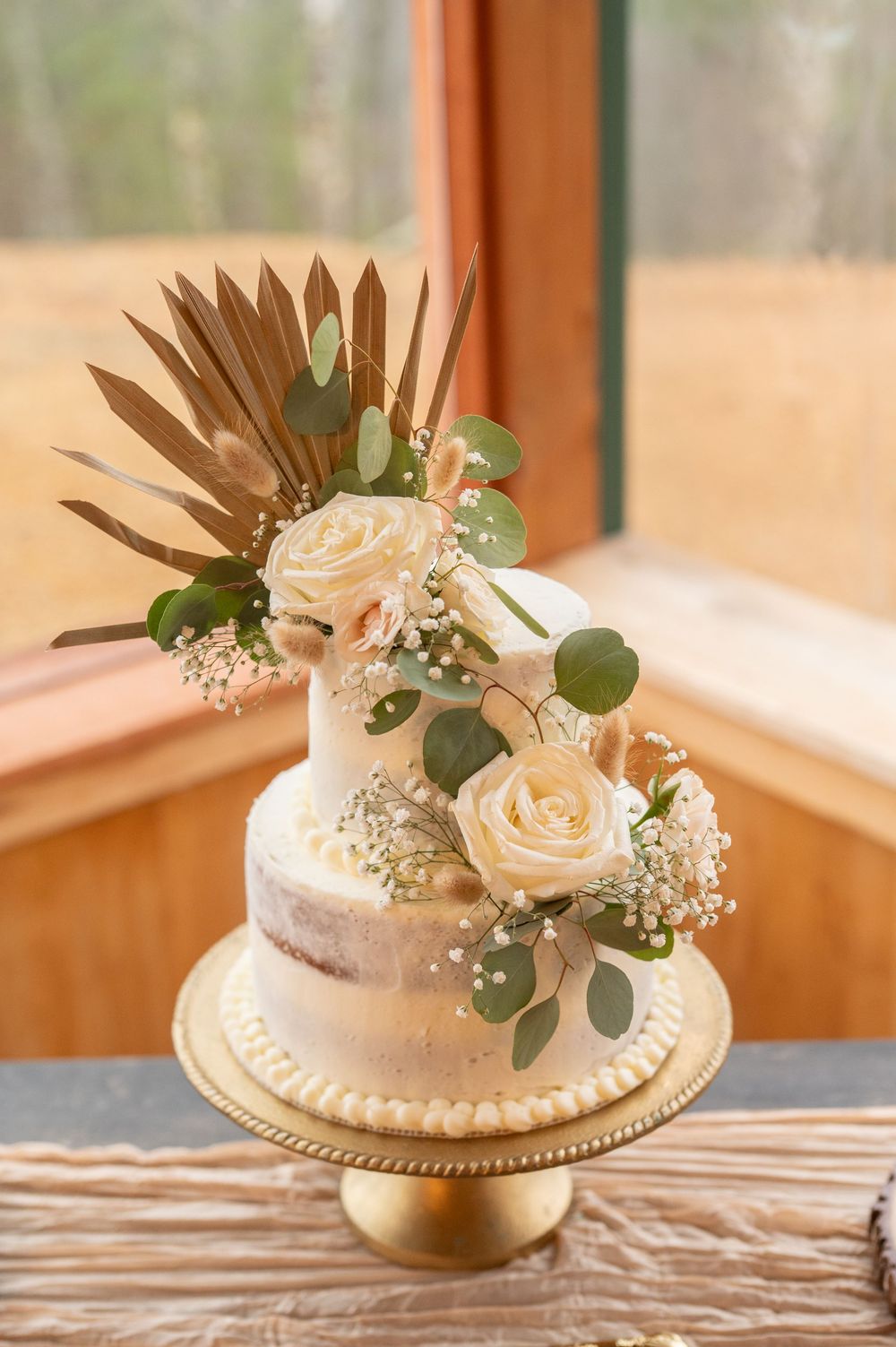 Wedding cake with peach roses and eucalyptus leaves on a white stand near a window in natural lighting.