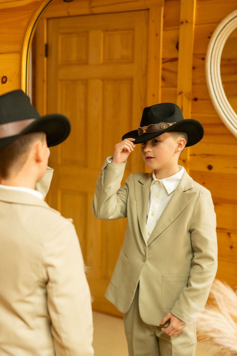 A young ring bearer in a beige suit and cowboy hat makes silly poses during a wedding photoshoot.