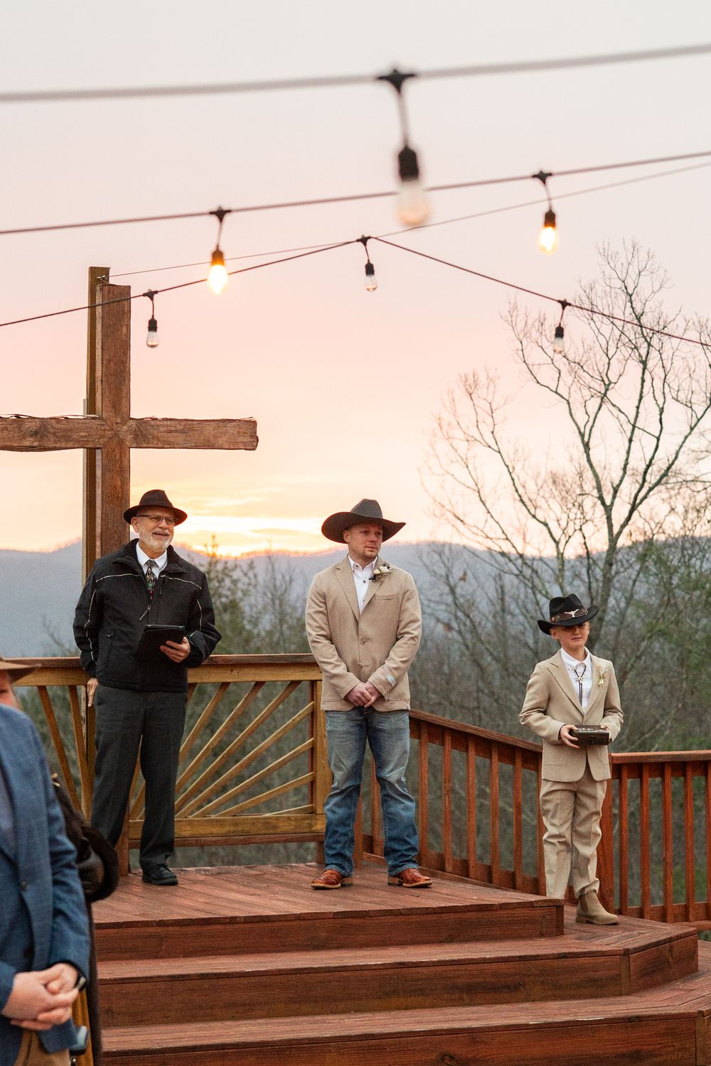 Three people in cowboy hats stand on wooden deck at sunset with string lights overhead and mountain views in background.