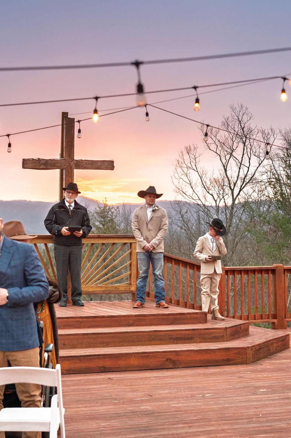 A rustic wooden deck with string lights and a cross during sunset at an outdoor ceremony in the mountains.