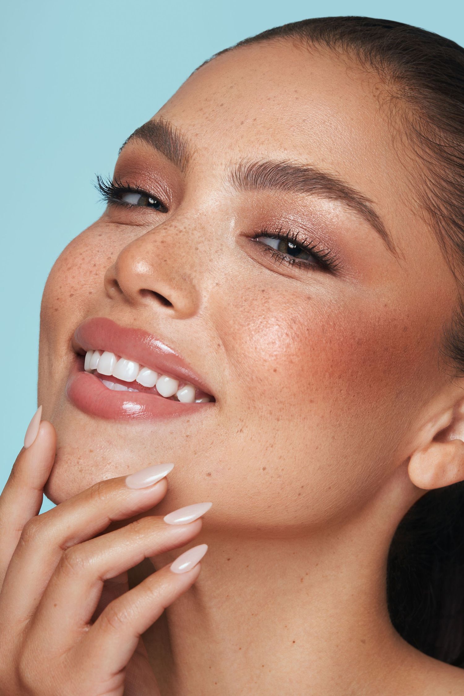 Close-up beauty portrait showing glowing skin, rosy makeup and perfect manicure against blue background.