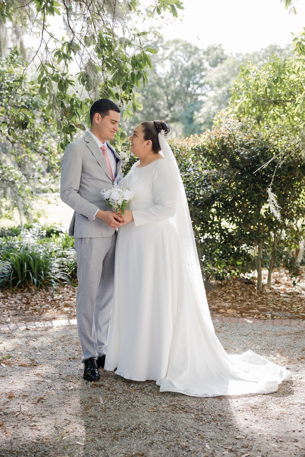 A bride and groom share a tender moment in a garden setting with sunlit trees and foliage in the background.