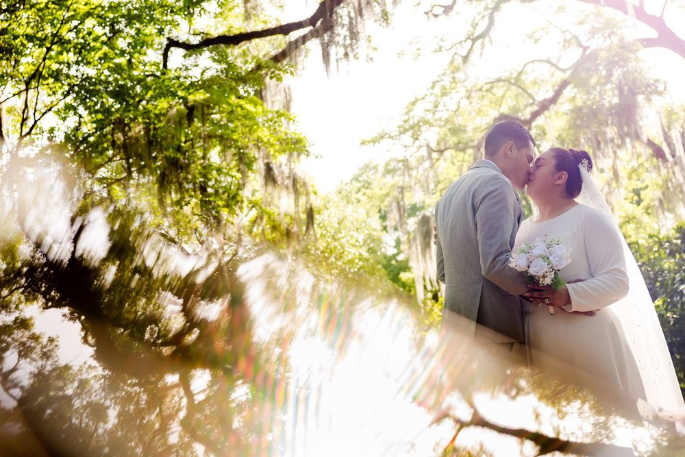 A romantic couple embraces outdoors in a sunlit garden with Spanish moss hanging from tree branches above.