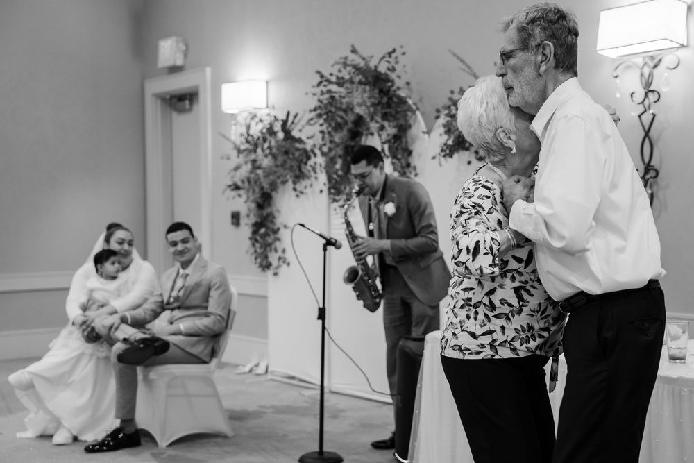 Black and white photo of wedding celebration with guests engaged in joyful conversation and laughter in a decorated hall.