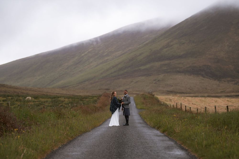 A couple walking down a misty rural road through rolling hills in the Scottish Highlands on an overcast day.