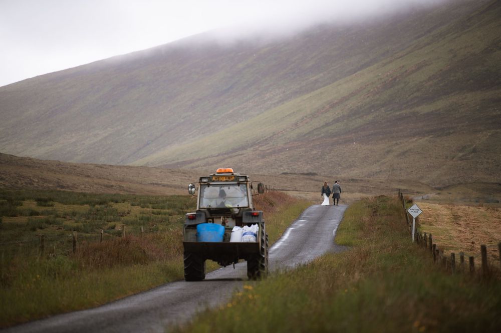 Blue tractor drives along a rural mountain road in misty Scottish highlands with rolling hills in the background.
