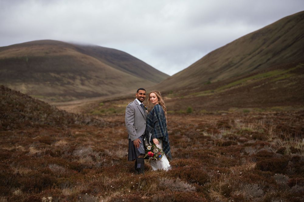 Two people stand together on a rugged hillside in the Scottish Highlands with brown hills and moody skies in the background.