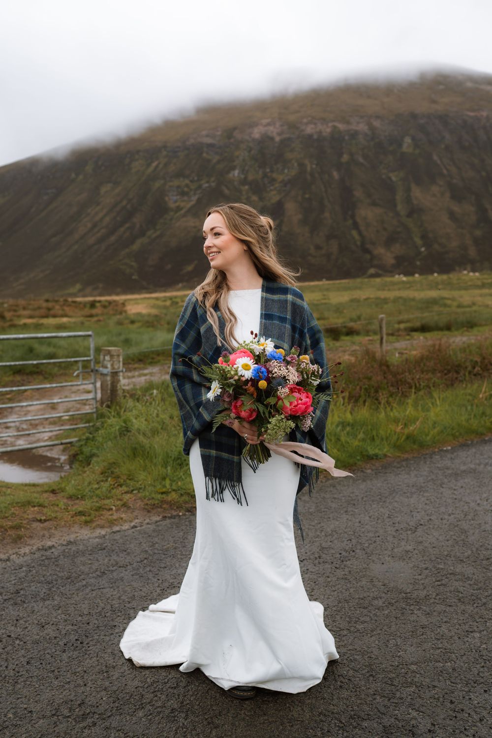 A bride in a white dress and plaid shawl holds colorful flowers on a misty mountain path in Scotland.