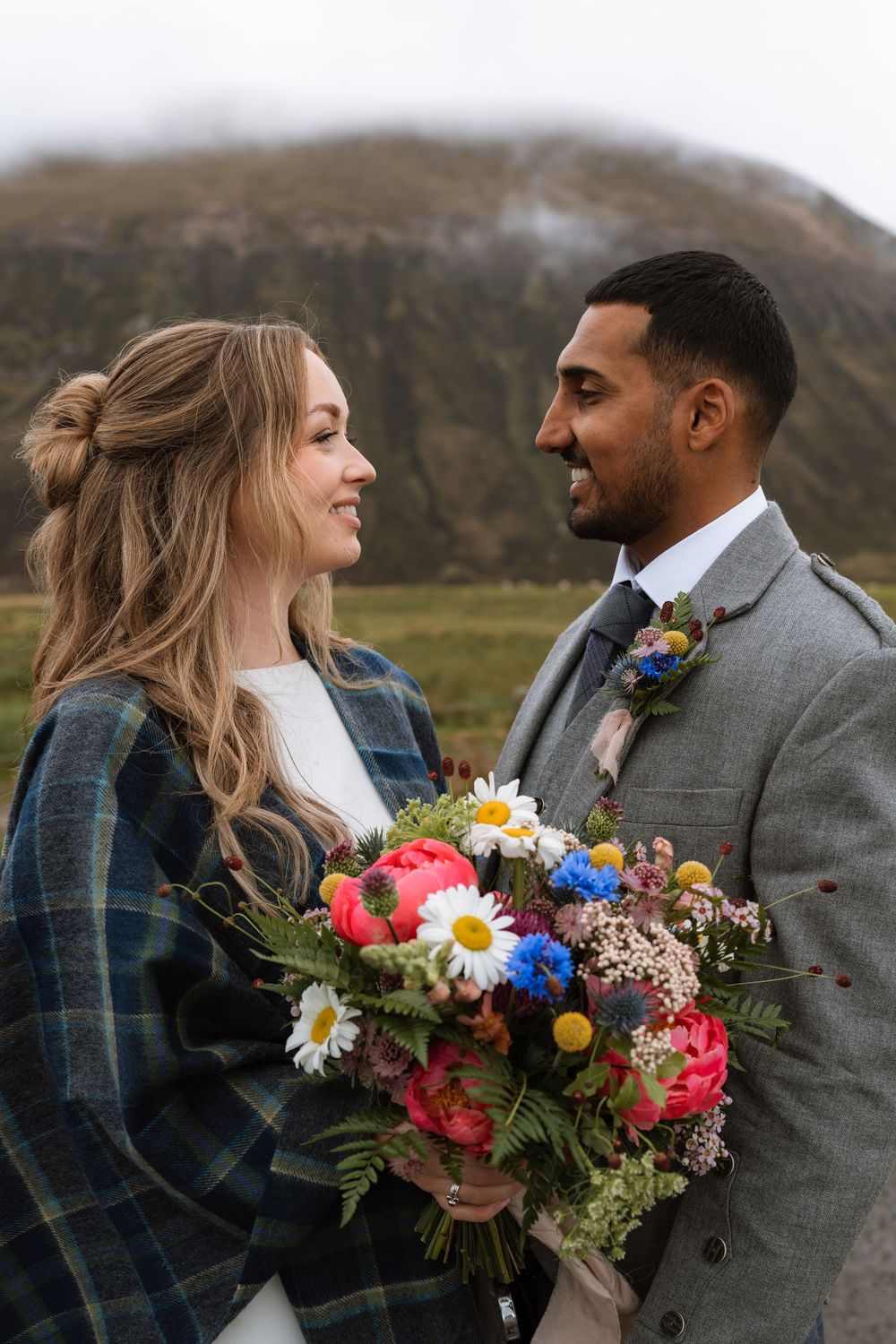 Two people in wedding attire smile at each other while holding a colorful bouquet in front of misty mountains in Scotland.