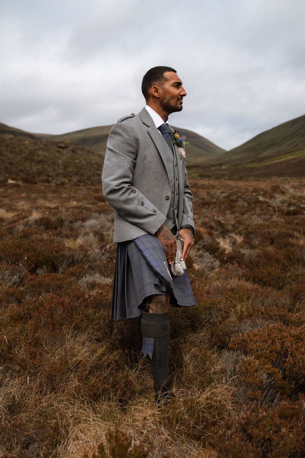 A person wearing a traditional Scottish kilt and gray suit jacket stands against a backdrop of rugged highland hills.