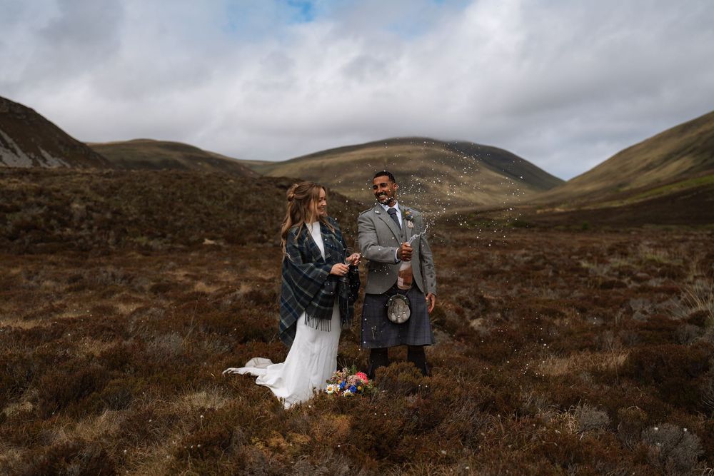 A couple in wedding attire stands together in the rugged Scottish Highlands beneath moody cloudy skies.
