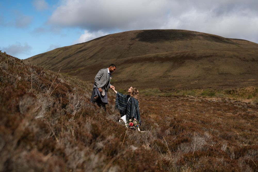 Two hikers traverse brown heathered slopes on a hillside in Scotland during an overcast day.