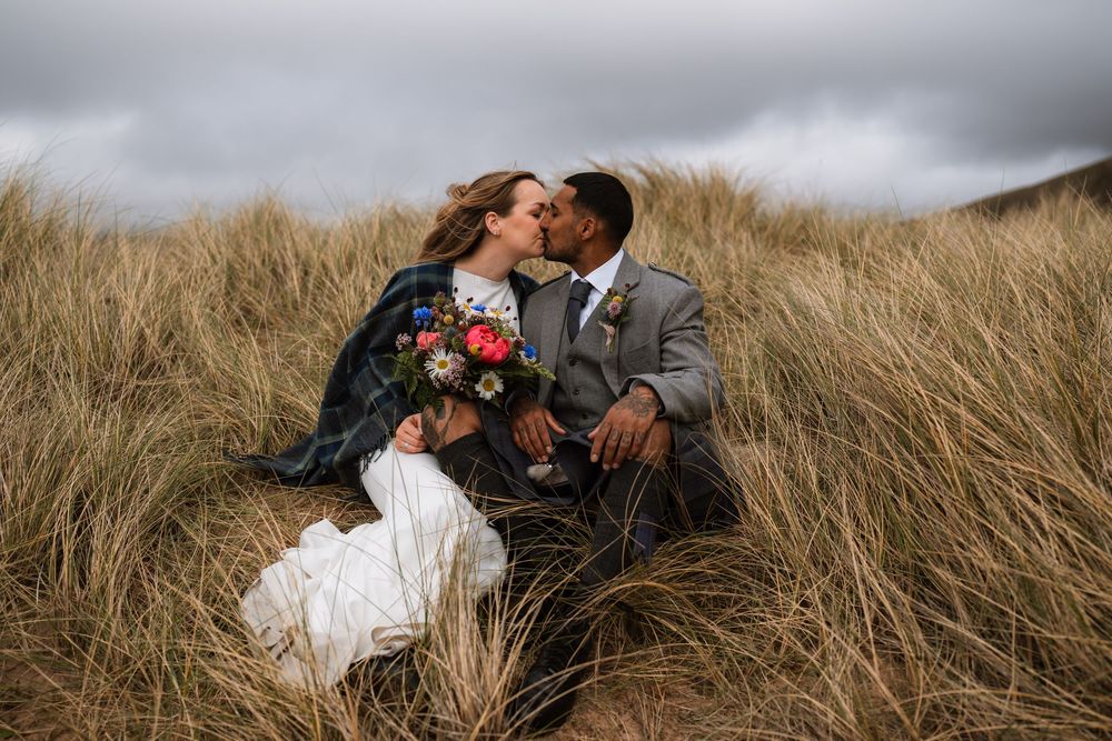 A couple sits intimately in tall golden grass against a moody sky while holding a floral bouquet.