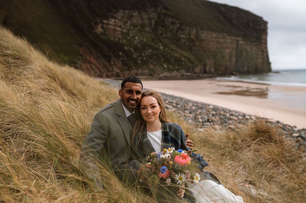 Couple snuggling on rocky coastal cliff with tall grass and ocean views during romantic outdoor photoshoot.
