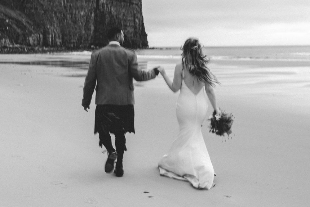 A couple walking along a misty beach shore holding hands in black and white photography.