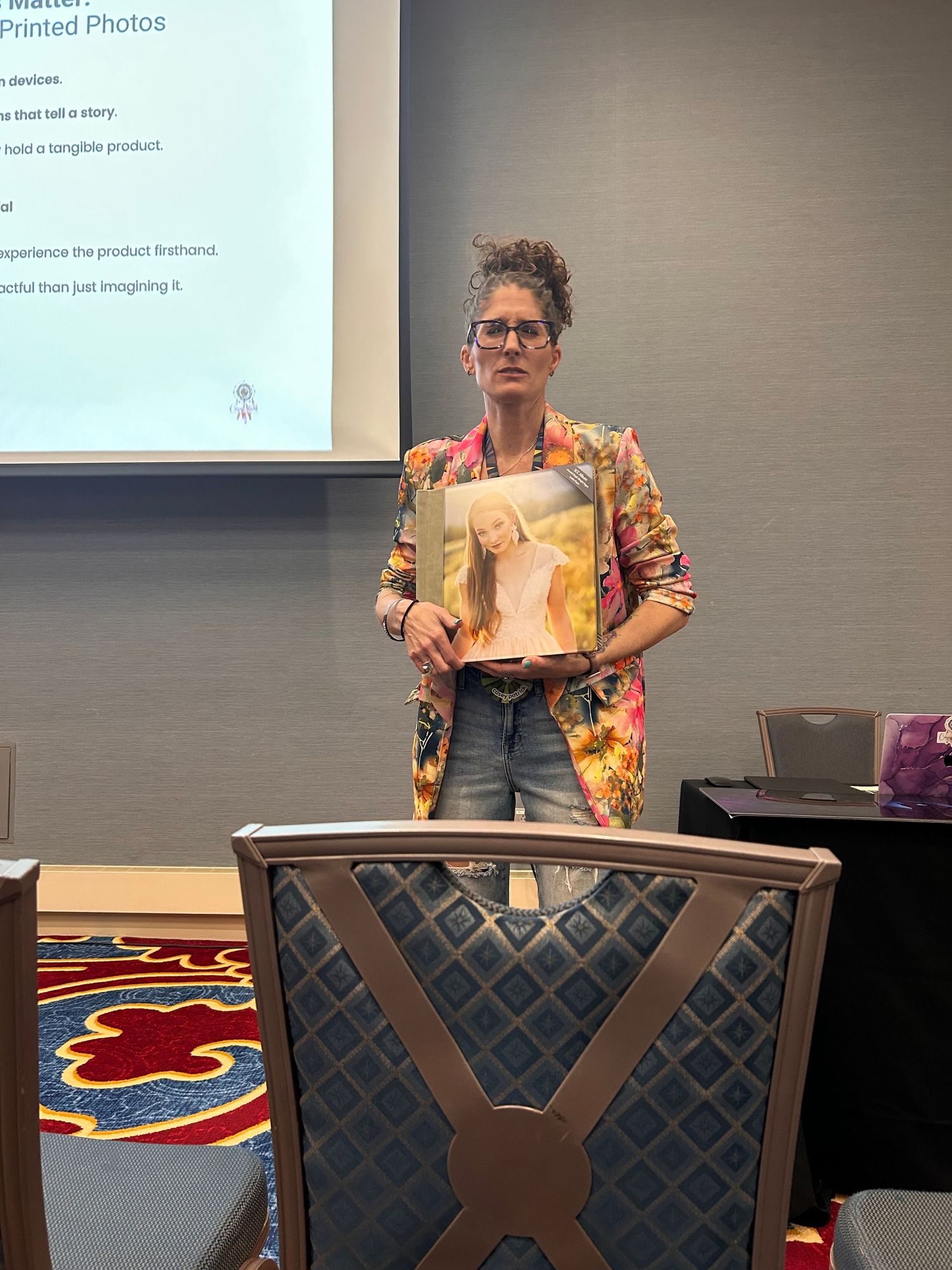 Presenter in a colorful floral blazer and glasses stands behind a chair while holding up a book in a classroom setting.