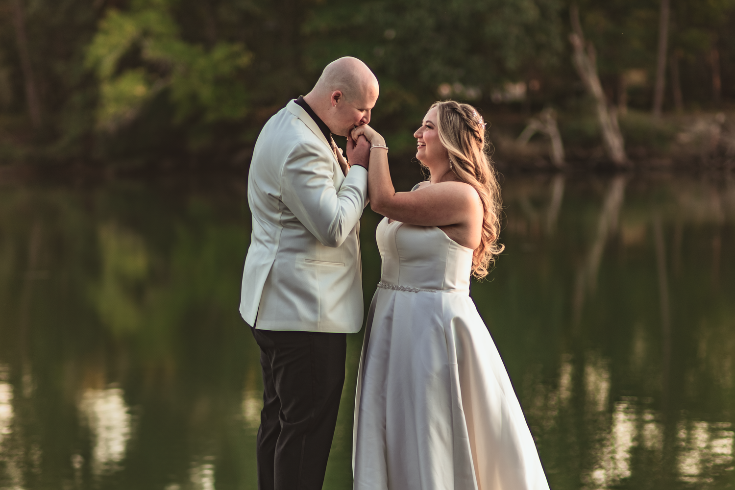 A couple shares a romantic embrace by a tranquil lake at sunset during an outdoor wedding ceremony.