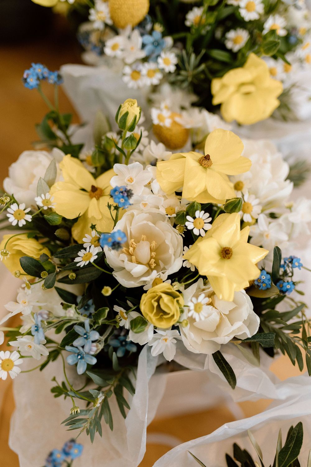 A delicate floral arrangement featuring yellow daffodils, white roses, and small blue forget-me-nots with green foliage.