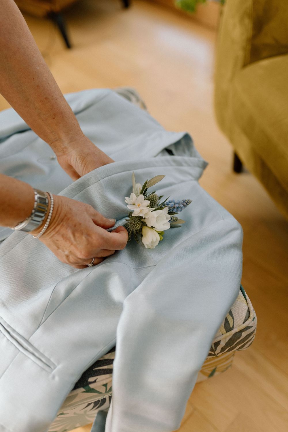 White boutonniere flower being pinned onto a light gray suit lapel in preparation for a formal event.
