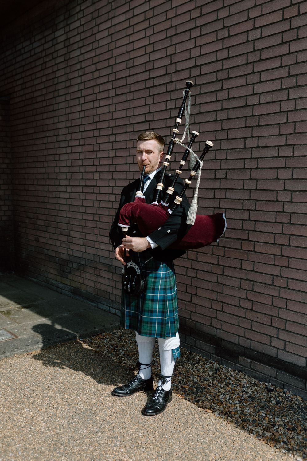 Bagpiper in traditional Scottish kilt and uniform playing instrument against brick wall in natural lighting.