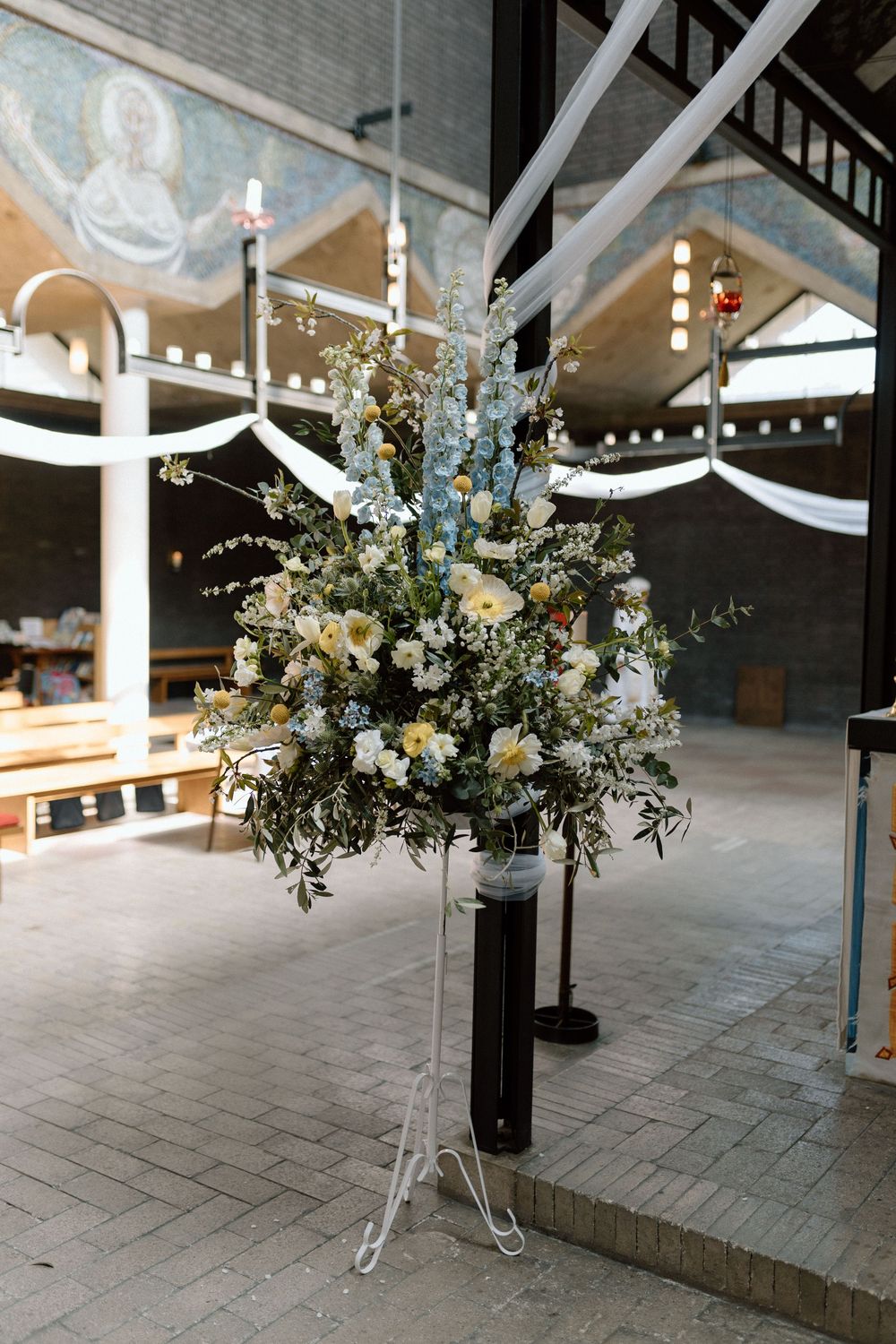 Elegant floral arrangement with white and yellow flowers on a vintage metal stand in an indoor venue with string lights.