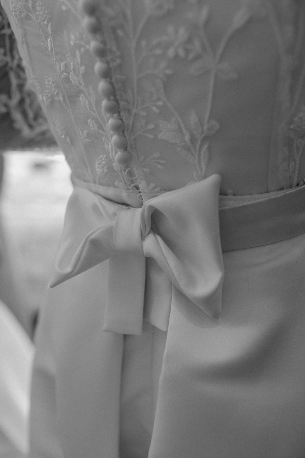 Black and white detail of an elegant bow tied at the back of a wedding dress with delicate lace embroidery detail.