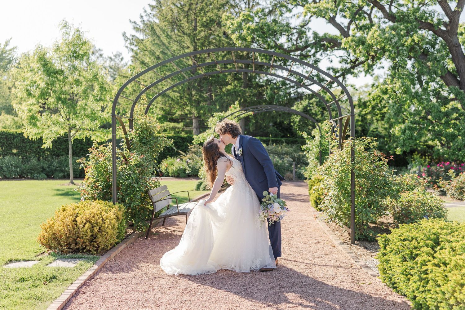 A couple shares a romantic moment under a garden arch surrounded by lush greenery and manicured landscaping.