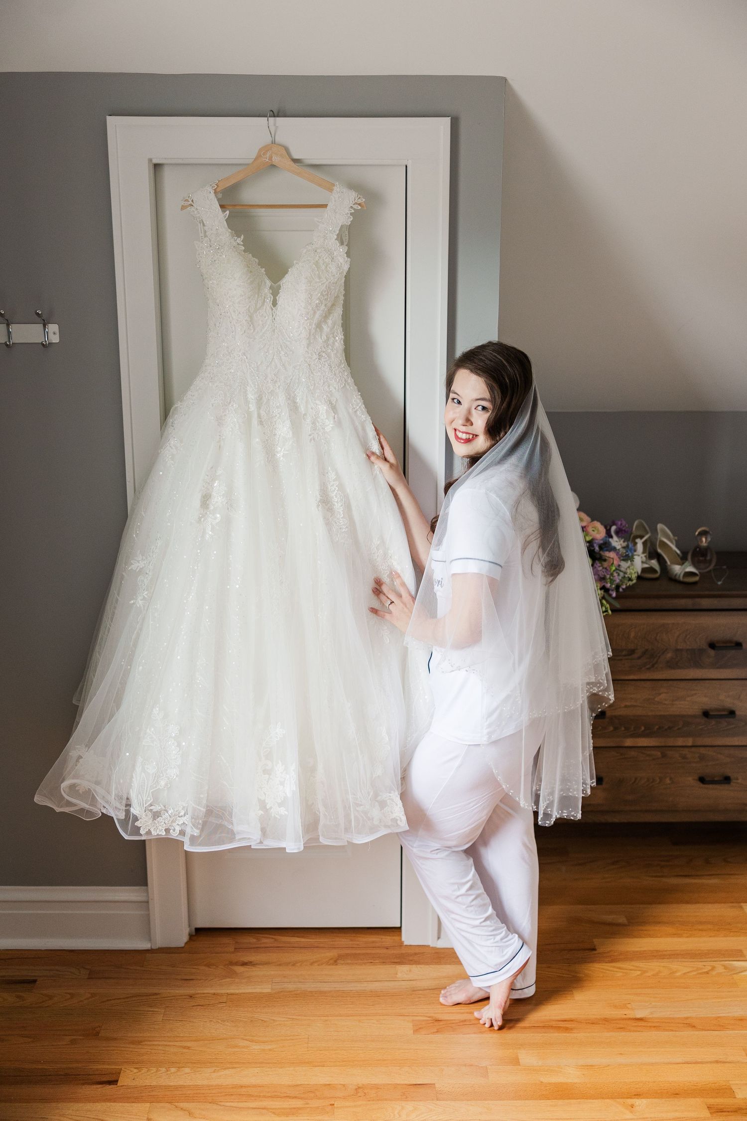 Elegant white lace wedding dress hangs on door while someone in white robe stands beside it excitedly.