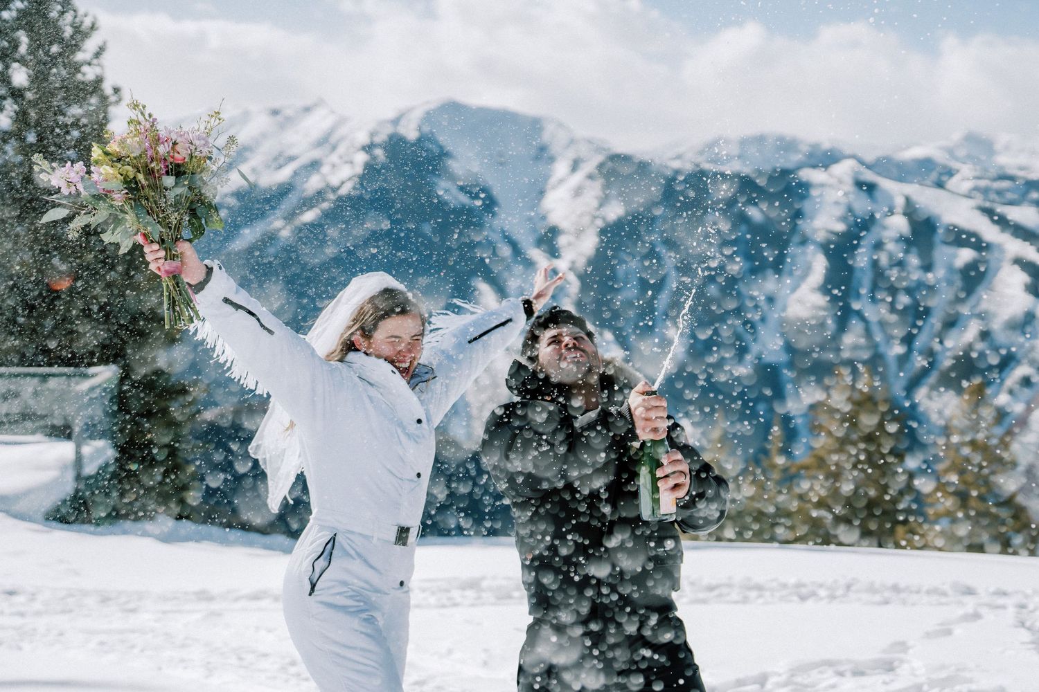Two people in winter clothing playing and throwing snow in front of snowy mountain landscape.