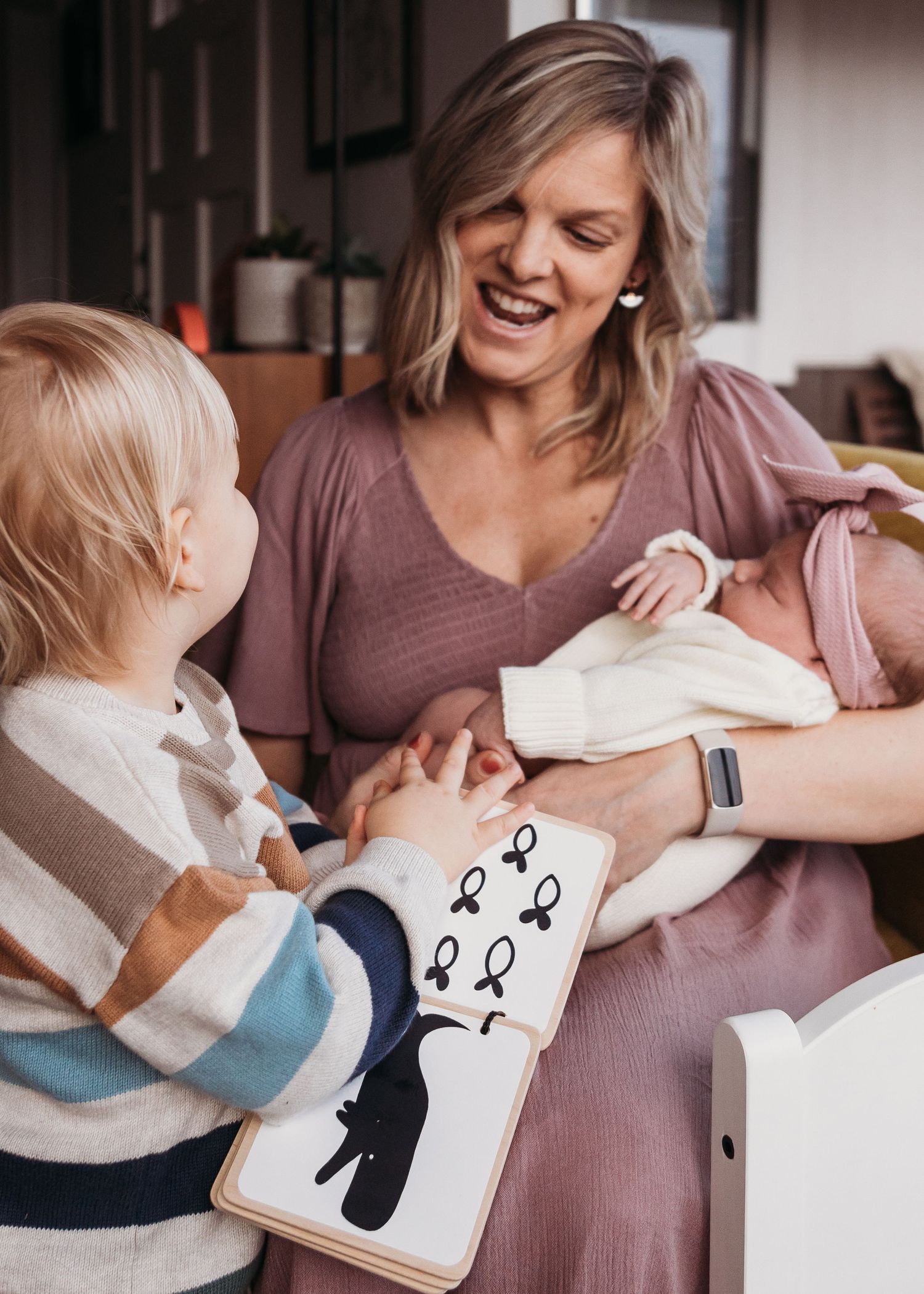 Candid family moment showing parent sharing a picture book with toddler while holding baby in cozy home setting.