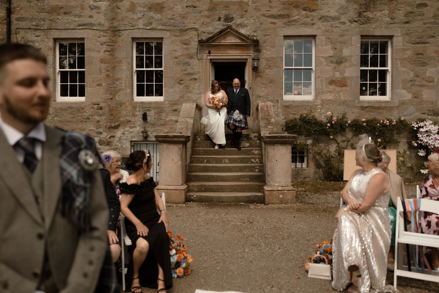 A cheerful wedding party celebrates on the grounds of a historic stone manor house in muted sepia tones.