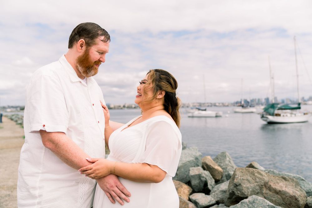 Loving couple embraces on rocky coastal shore with boats and harbor visible in background on cloudy day.