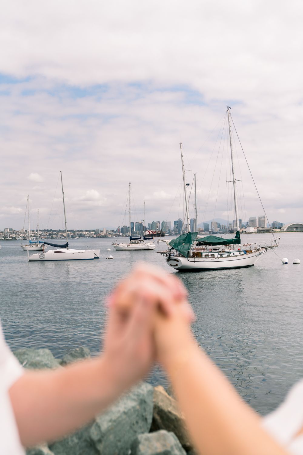 Hands clasped together overlooking a marina with sailboats and city skyline in the background under cloudy skies.