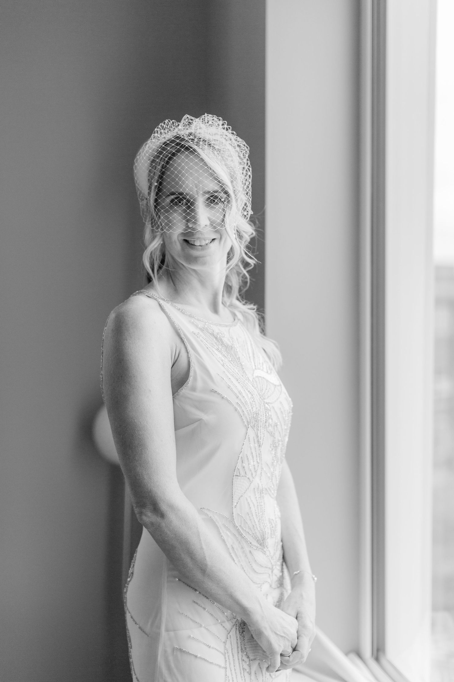 Black and white portrait of a bride wearing a vintage-inspired lace dress and birdcage veil standing by a window in soft light cotswolds