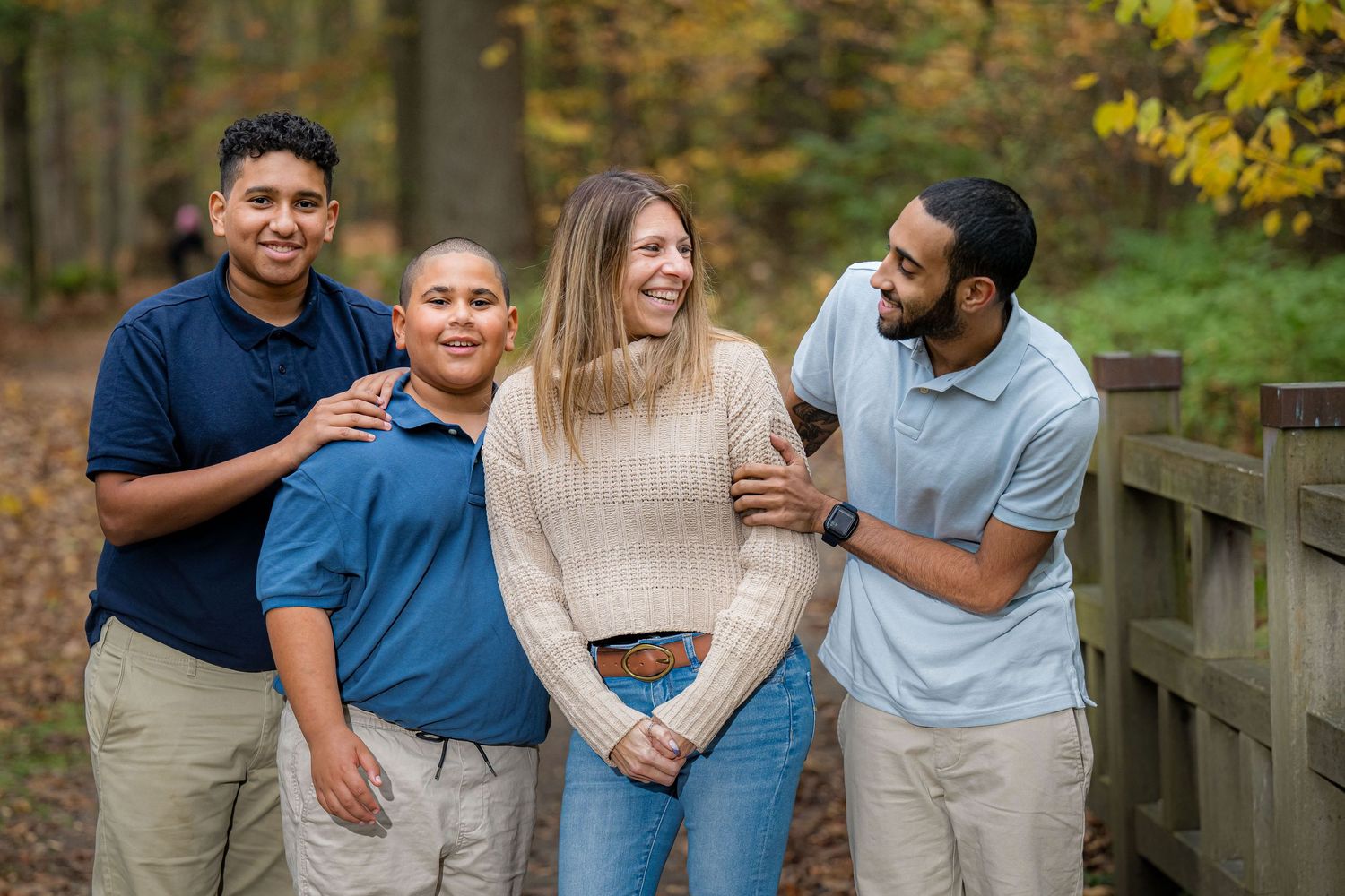 A group laughs together while standing on a wooden bridge surrounded by autumn foliage in a park setting.