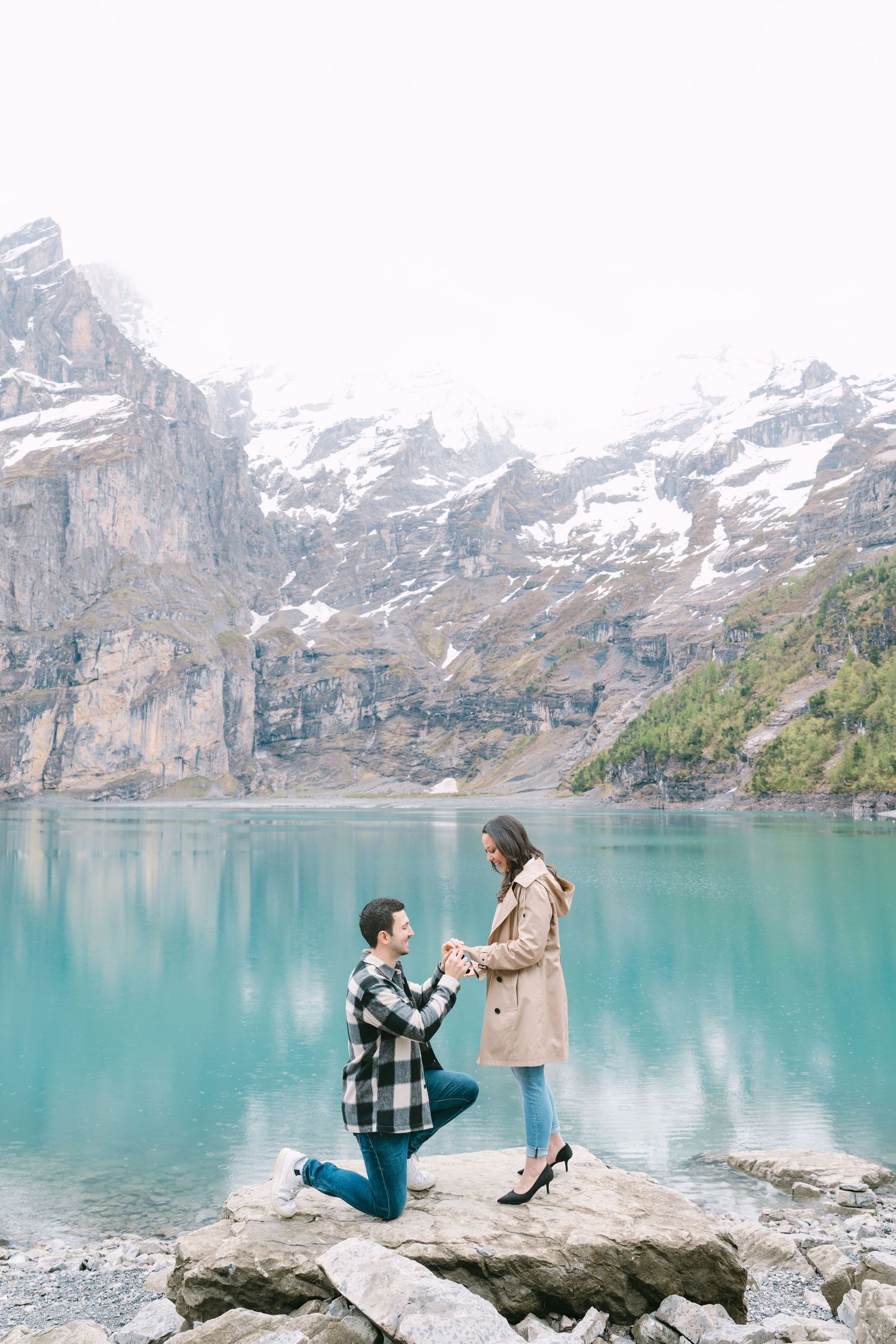 A couple shares an intimate moment during a marriage proposal by a turquoise alpine lake with snow-capped mountains in background.