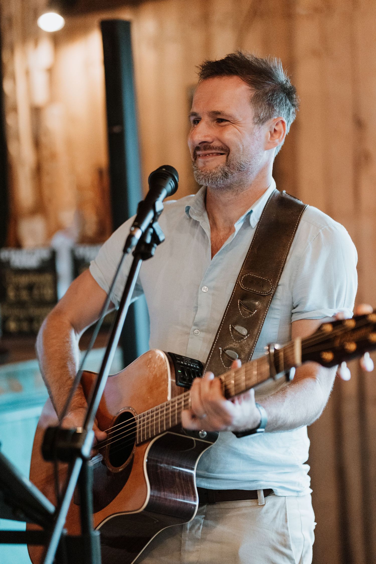 Guitarist in white shirt performs on acoustic guitar with microphone at rustic wood-paneled venue in warm lighting.