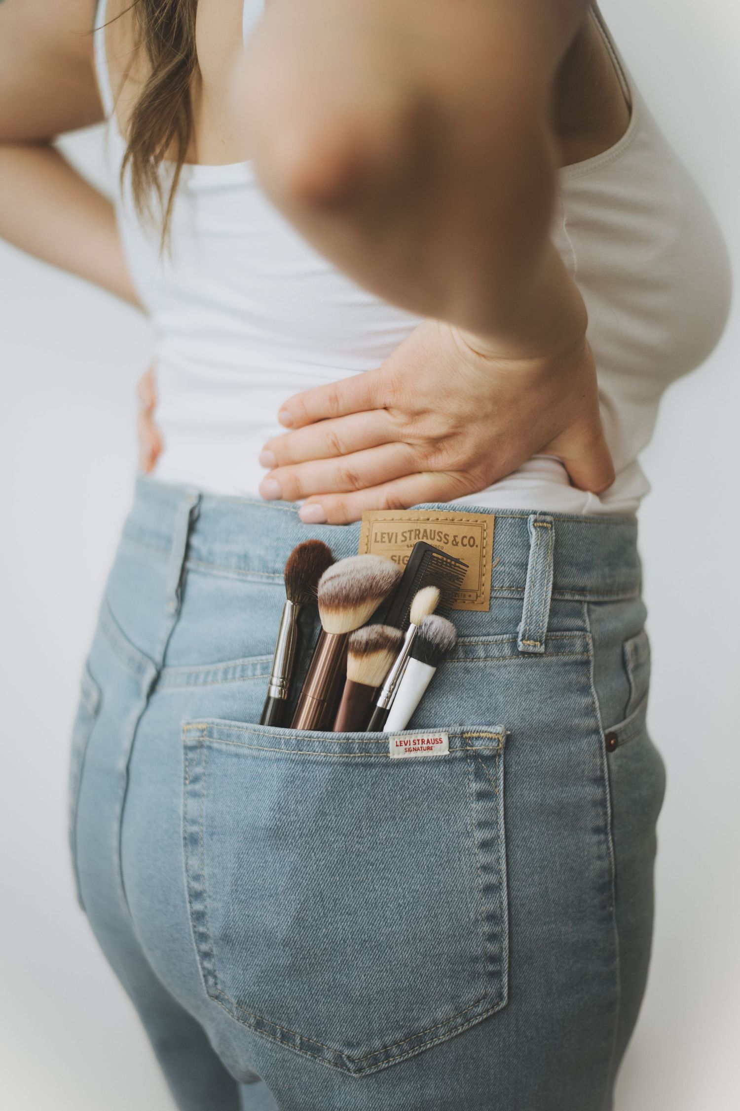 Makeup brushes tucked into the back pocket of blue jeans against a white background.