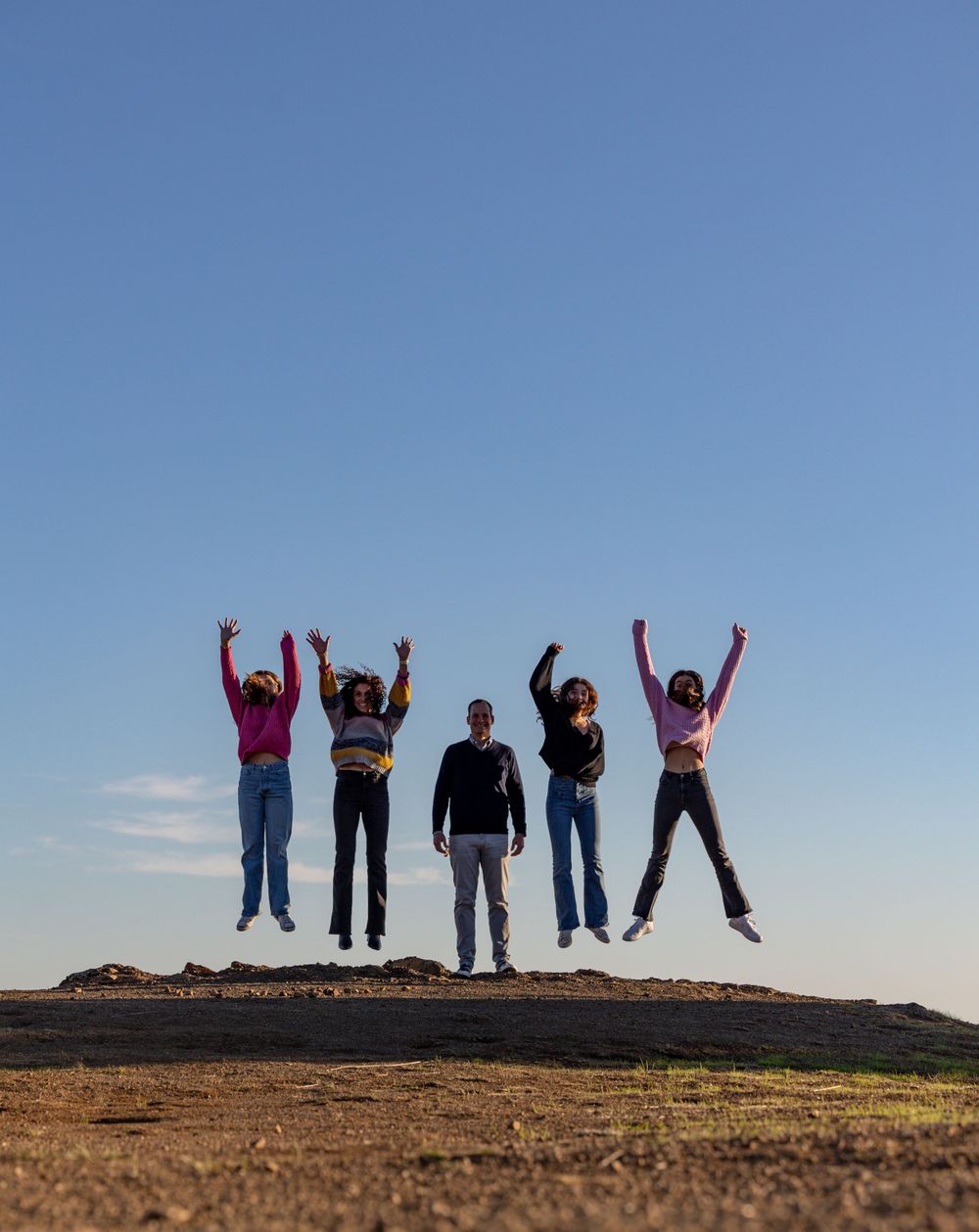 A group of people jumping in unison against a blue sky on a hilltop during sunset.