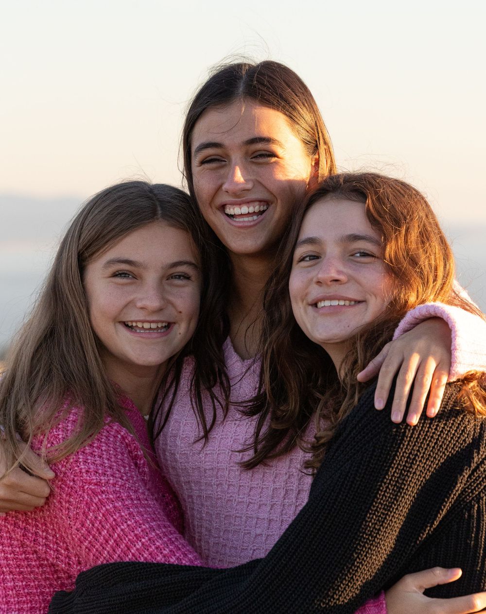 Three friends wearing pink and brown sweaters embrace and smile warmly during golden hour.