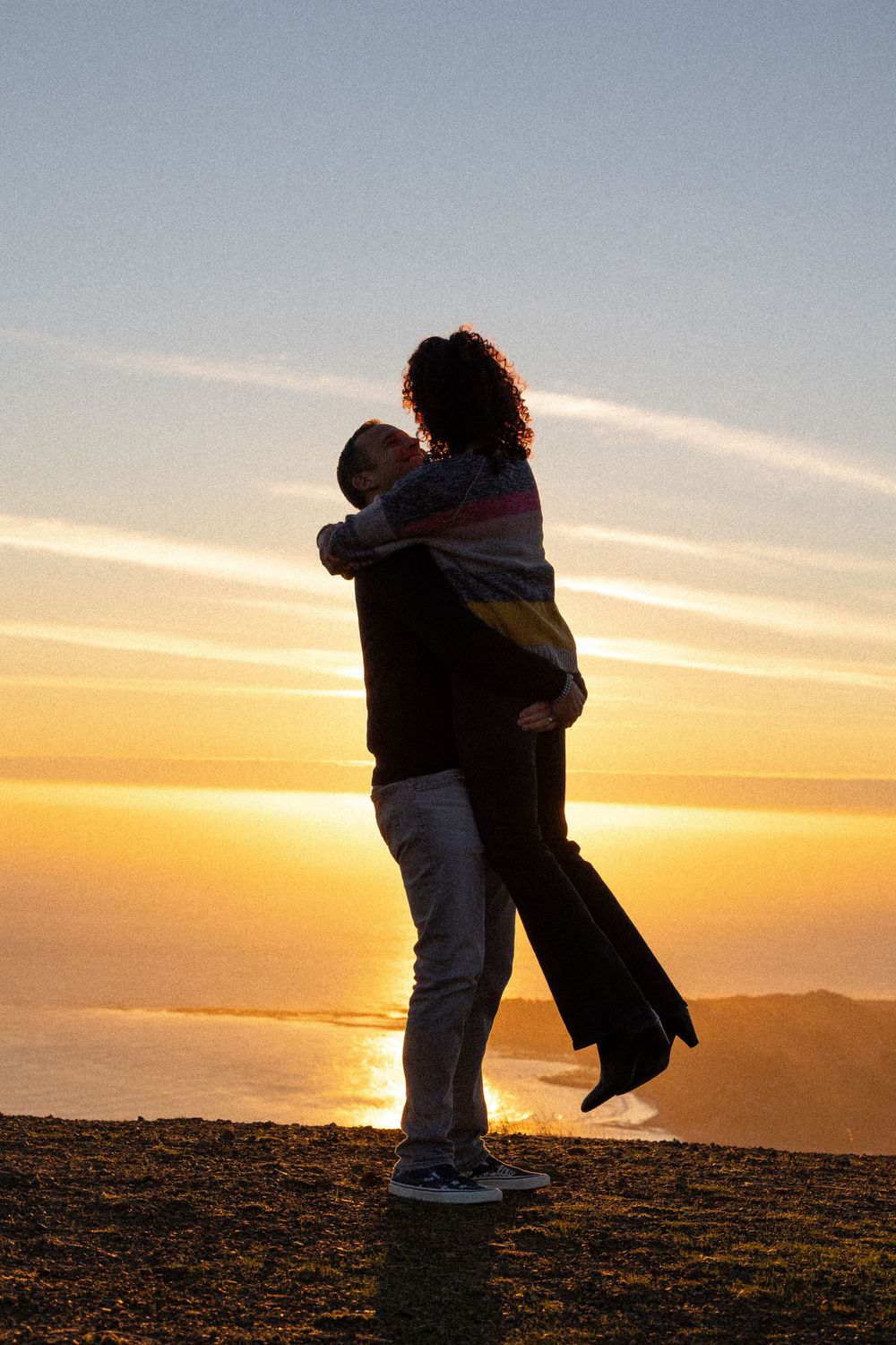Silhouetted couple embrace during golden sunset over mountain landscape as sun touches horizon.
