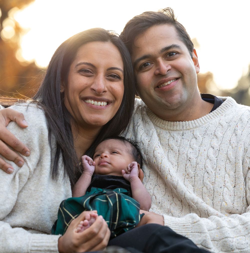 Couple in white sweaters hold their sleeping newborn baby wearing green during a warm sunset outdoors.