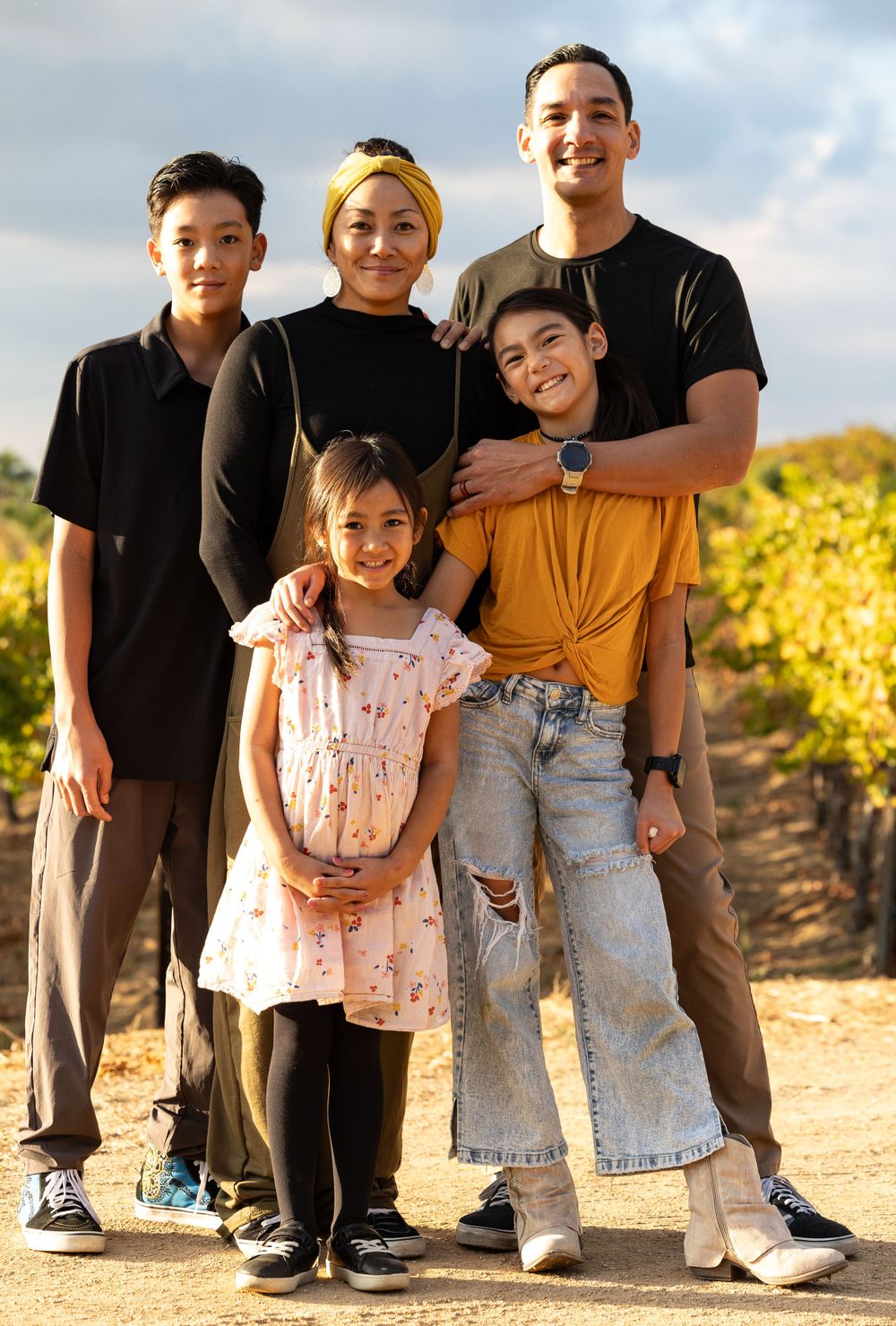 A family of five poses together outdoors on a dirt path with yellow foliage in the background on a sunny day.