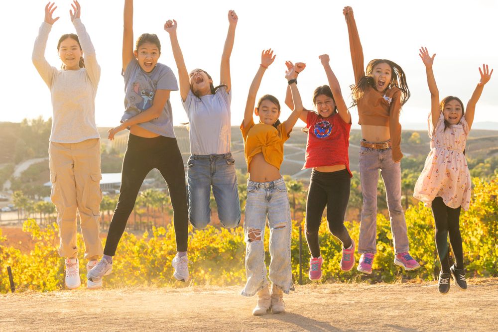 Group of children jumping with joy together in a field during golden hour with vineyards in the background.