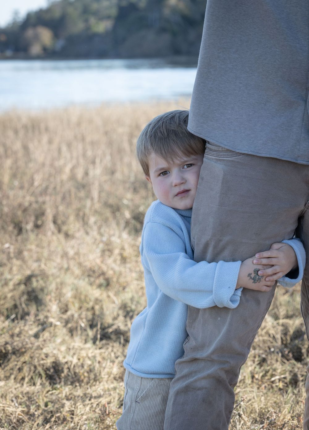 A small child in a light blue sweater hugs onto an adult's leg in a grassy field by the water.