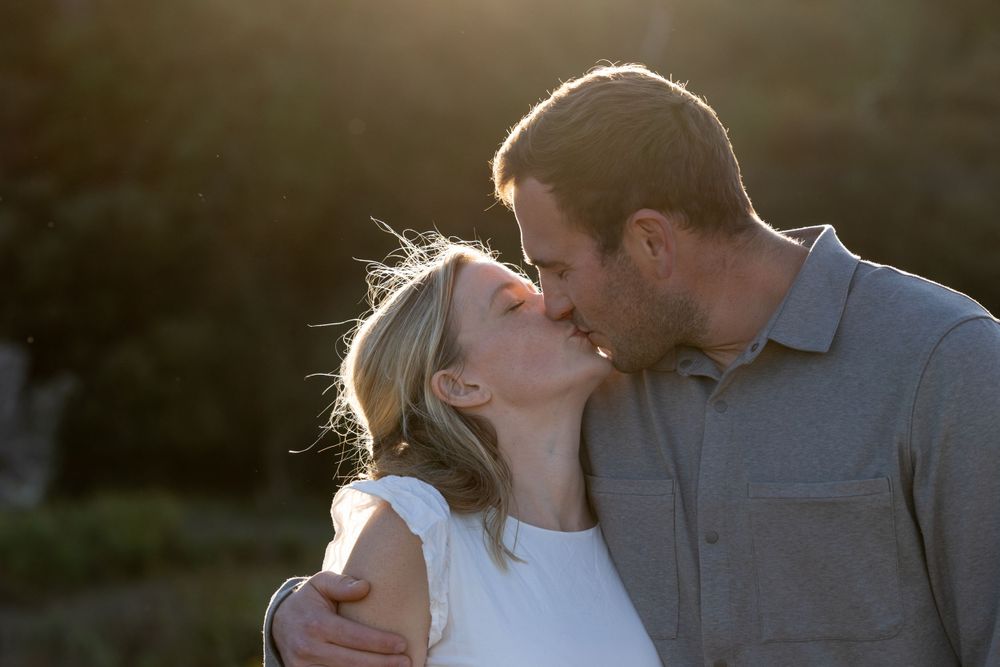 A couple shares a tender kiss outdoors during golden hour with sunlit backlighting creating a romantic silhouette.
