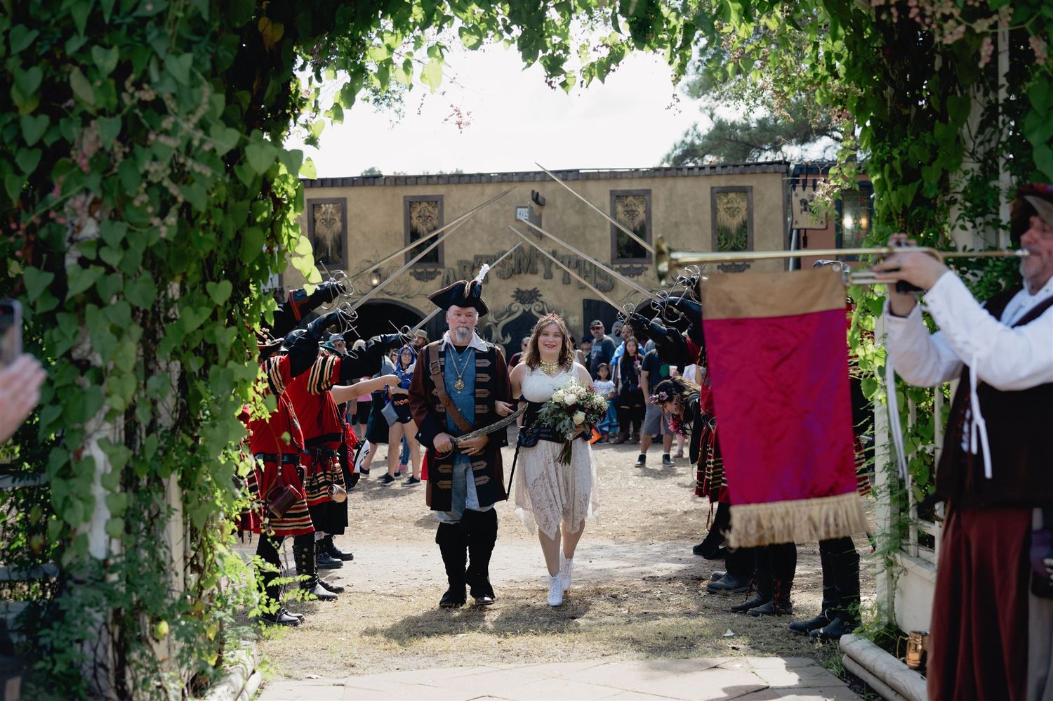 A wedding ceremony takes place outdoors under a lush green archway as guests gather to celebrate.
