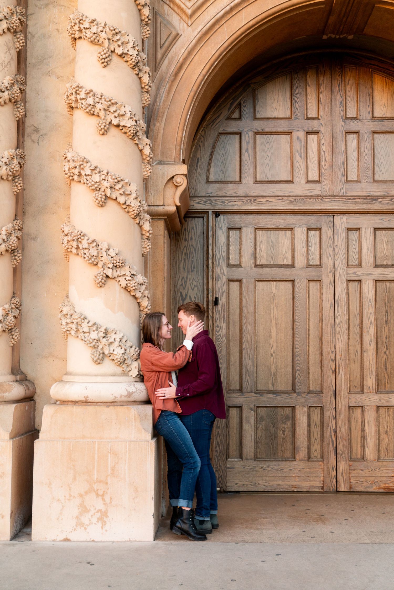 A couple shares an intimate moment against an ornate stone column and wooden door at a historic building entrance.