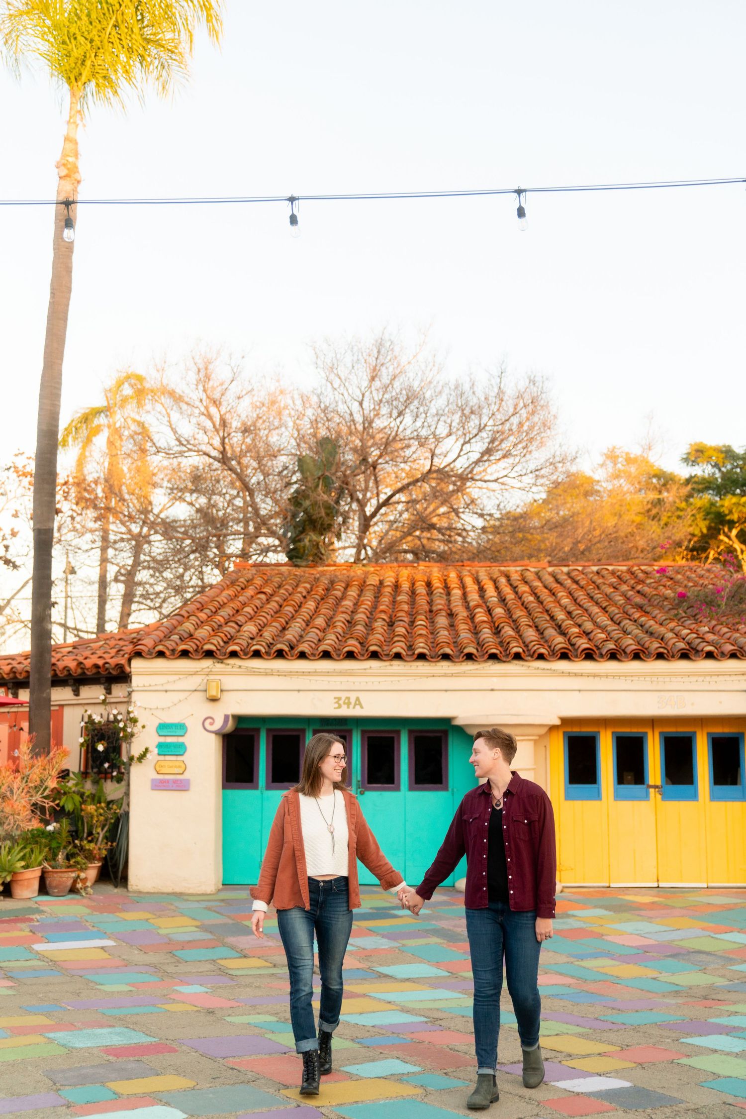 Couple walking hand in hand on colorful mosaic courtyard with Spanish-style building and palm trees in warm sunset light.